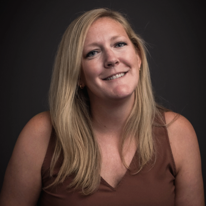 A woman with long blonde hair, wearing a sleeveless brown top, smiles slightly while posing against a dark gray background, embodying the confidence and stability often associated with Permanent Equity.