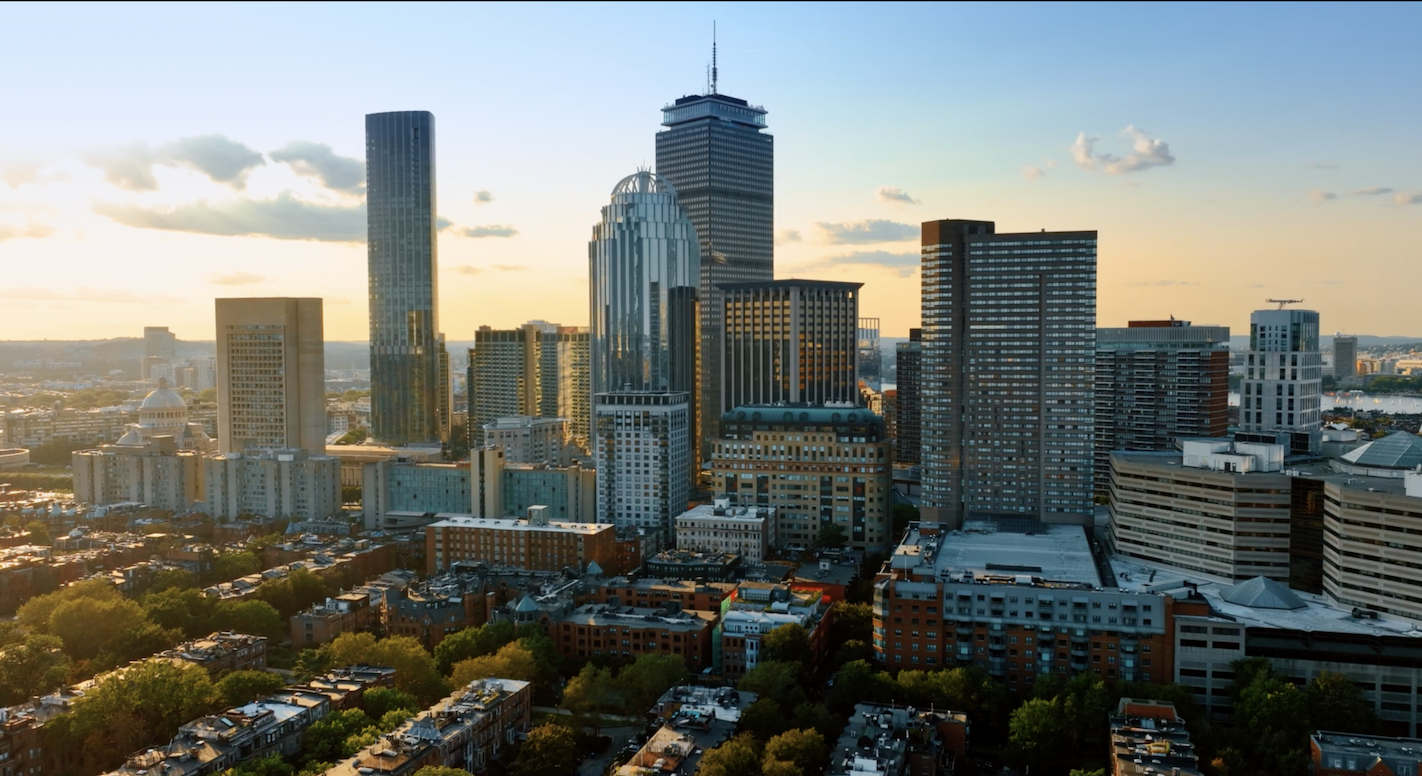 A city skyline at sunset with tall modern skyscrapers, mid-rise buildings, and green trees in the foreground under a clear sky with scattered clouds.