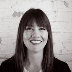 Morgan Witham, a woman with straight, dark hair and bangs, smiles at the camera while standing in front of a white brick wall. The image is in black and white.