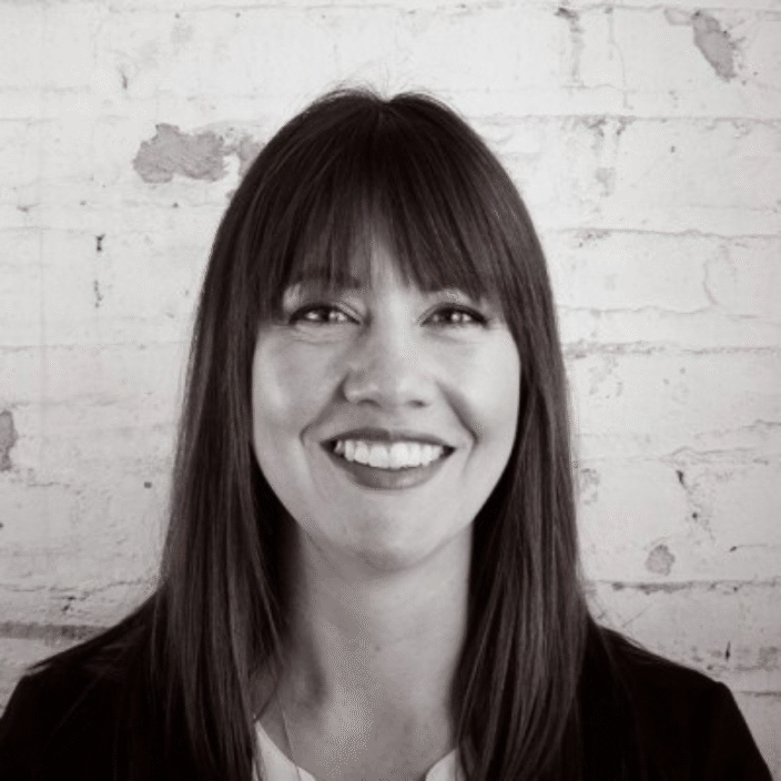Morgan Witham, a woman with straight, dark hair and bangs, smiles at the camera while standing in front of a white brick wall. The image is in black and white.