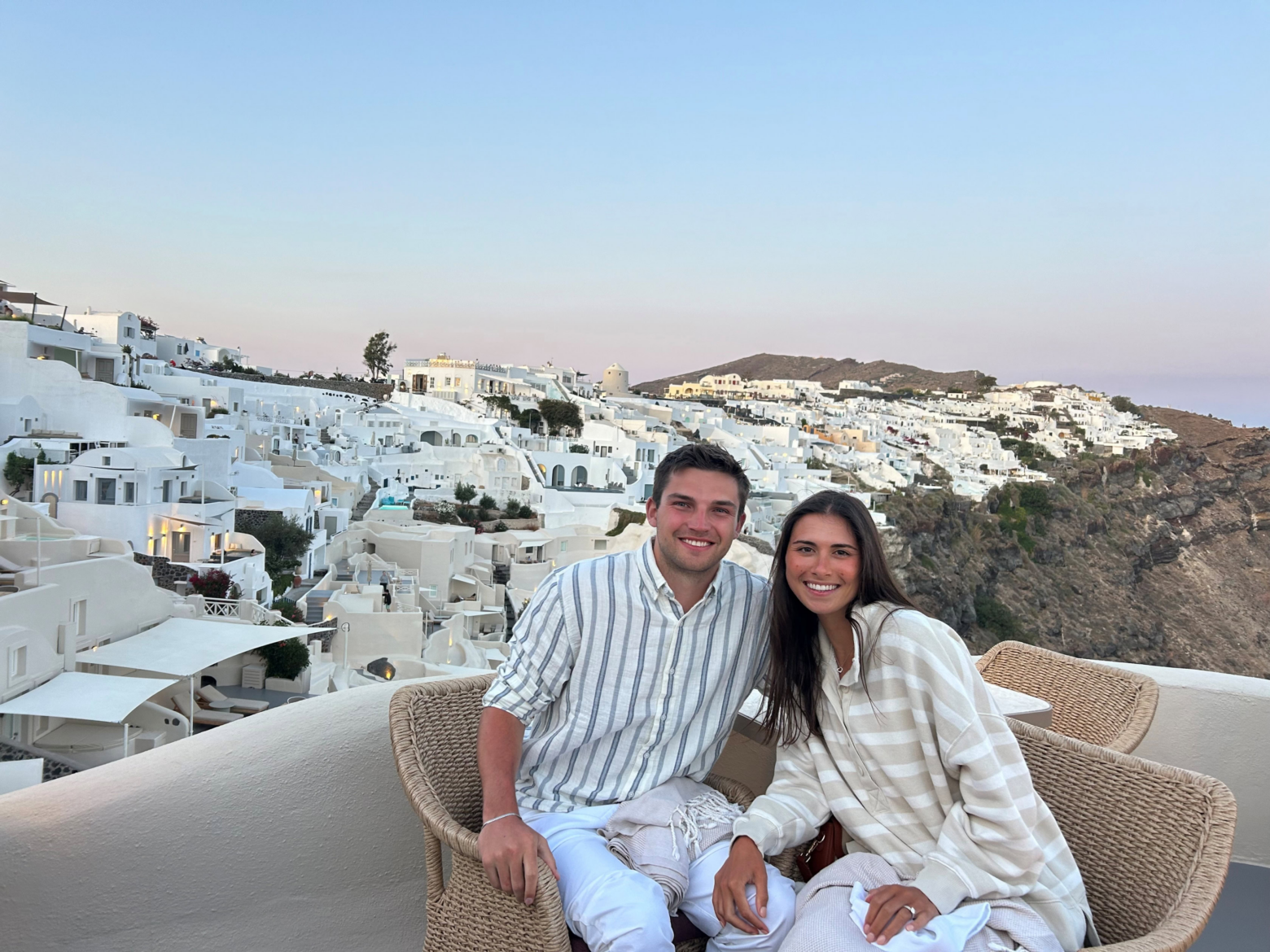 A smiling couple in light-colored clothes sits on wicker chairs, surrounded by white buildings on a hillside in Santorini, Greece, during sunset—capturing the serene beauty often seen in Kevin Corcoran’s picturesque photography.