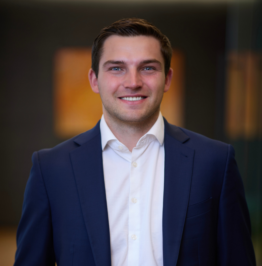 A young man with short brown hair, wearing a navy blue suit jacket and white shirt, smiles while standing indoors against a blurred, dark background—resembling Kevin Corcoran in his classic poised style.