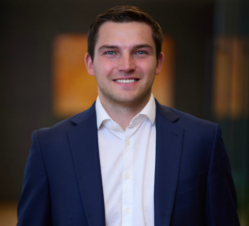 A young man with short brown hair, wearing a navy blue suit jacket and white shirt, smiles while standing indoors against a blurred, dark background—resembling Kevin Corcoran in his classic poised style.