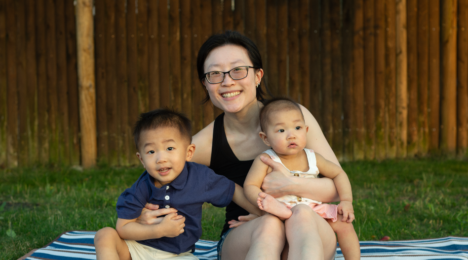 A smiling woman with glasses sits on a striped blanket outdoors, holding a baby on her lap and embracing a young boy beside her. They are in front of a wooden fence on grass.