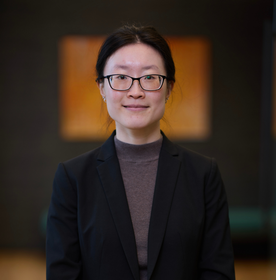 A woman with glasses and long dark hair tied back, wearing a dark blazer and brown top, stands indoors in front of a blurred background with warm-toned artwork.