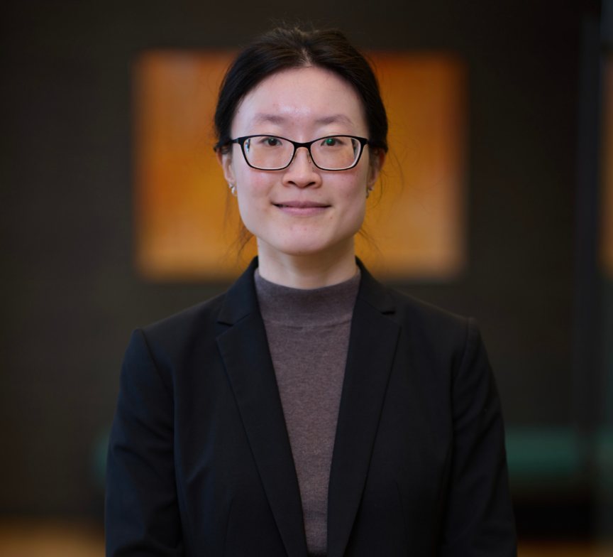 A woman with glasses and long dark hair tied back, wearing a dark blazer and brown top, stands indoors in front of a blurred background with warm-toned artwork.