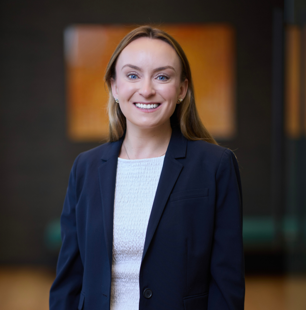 A woman with long light brown hair, wearing a navy blazer and white top, smiles at the camera in a softly lit indoor setting with a blurred background.