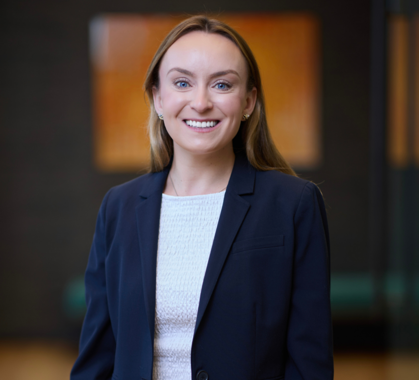 A woman with long light brown hair, wearing a navy blazer and white top, smiles at the camera in a softly lit indoor setting with a blurred background.