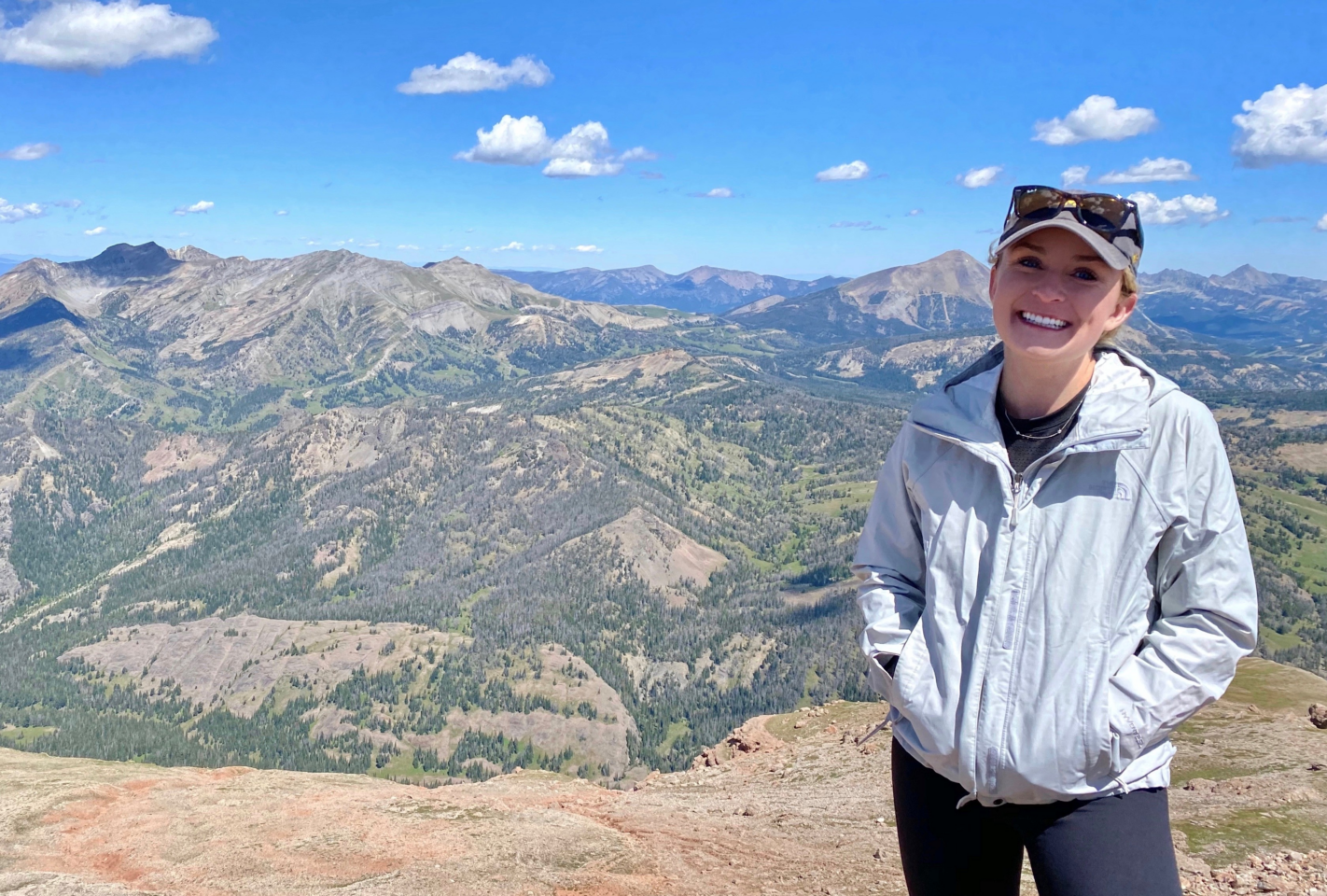Anna Hilzinger, in a light jacket and sunglasses, stands smiling on a mountain summit, with expansive mountain ranges and a blue sky with scattered clouds in the background.