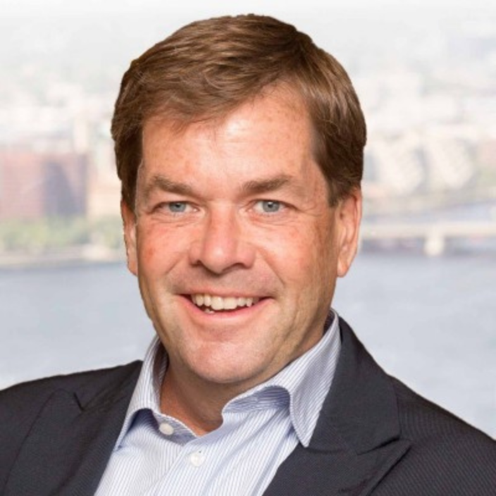 A man with short brown hair, wearing a dark blazer and light blue collared shirt, is smiling at the camera with a blurred cityscape and river in the background, reflecting his professional role at Bain Capital.