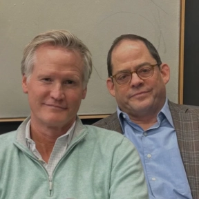 Two middle-aged men pose together indoors for a No Guest Episode. One wears a light green pullover over a collared shirt, while the other sports glasses, a blue shirt, and a plaid blazer. A light-colored wall serves as the background.