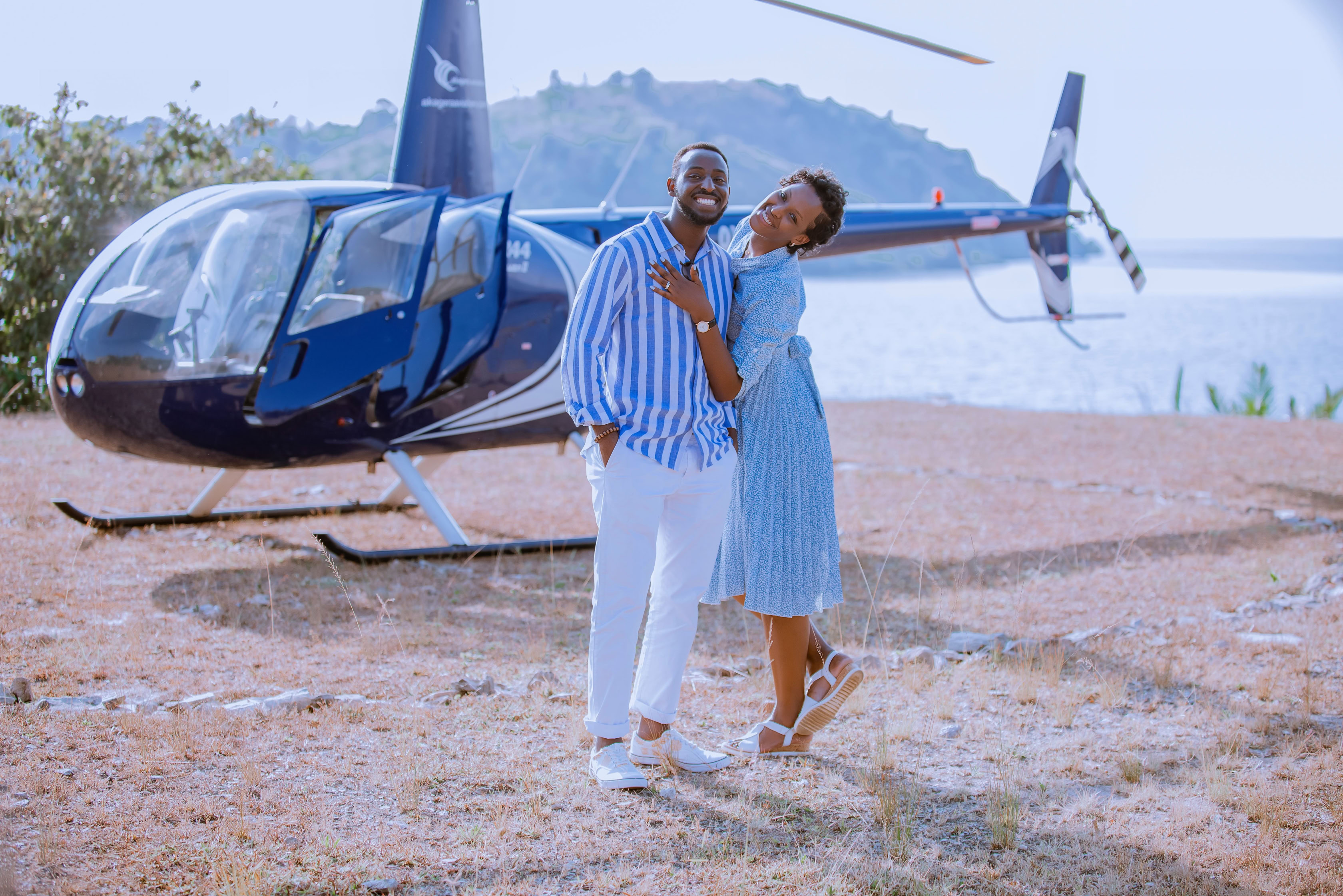 A smiling couple stands together in front of a blue helicopter near a body of water, both wearing blue and white outfits on a sunny day, reminiscent of the adventurous spirit often captured by Irénée Zigururimi.