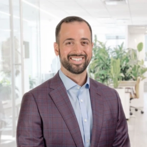 Mike Alberts, a man with short dark hair and a beard, wearing a purple plaid suit jacket and blue shirt, smiles while standing in a bright, modern office with plants and glass walls in the background.
