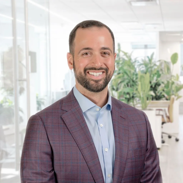 Mike Alberts, a man with short dark hair and a beard, wearing a purple plaid suit jacket and blue shirt, smiles while standing in a bright, modern office with plants and glass walls in the background.