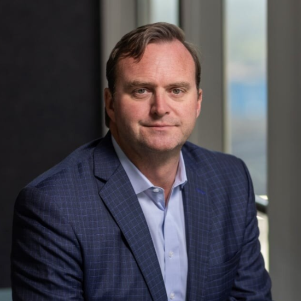 A man with short brown hair wearing a blue checked suit jacket and light blue shirt sits in front of large windows, looking at the camera with a neutral expression—Chuck Thompson exudes quiet confidence.